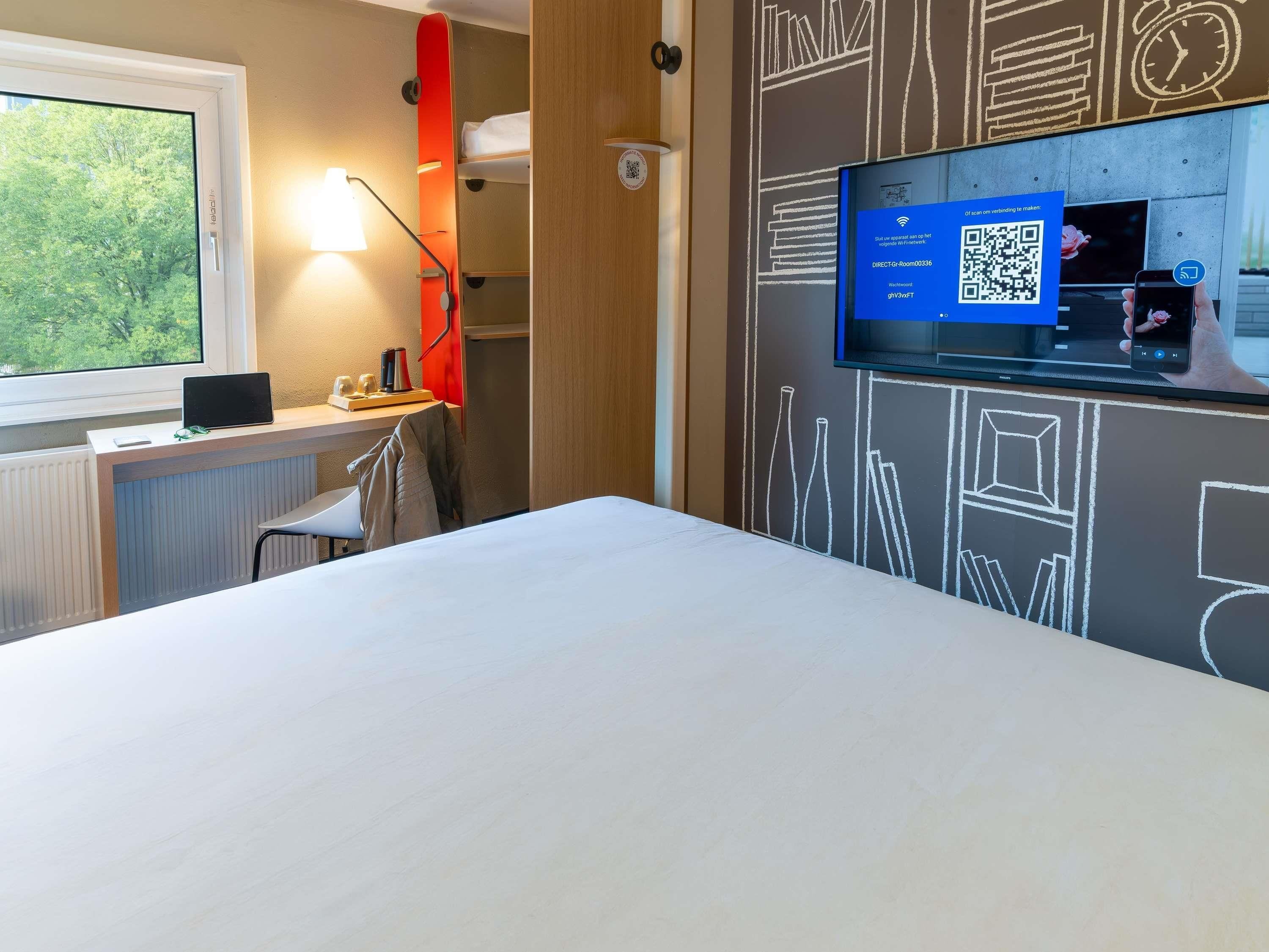 Een hotelkamer met een bed, een bureau met een laptop, een aan de muur bevestigde televisie en een raam met uitzicht op bomen.