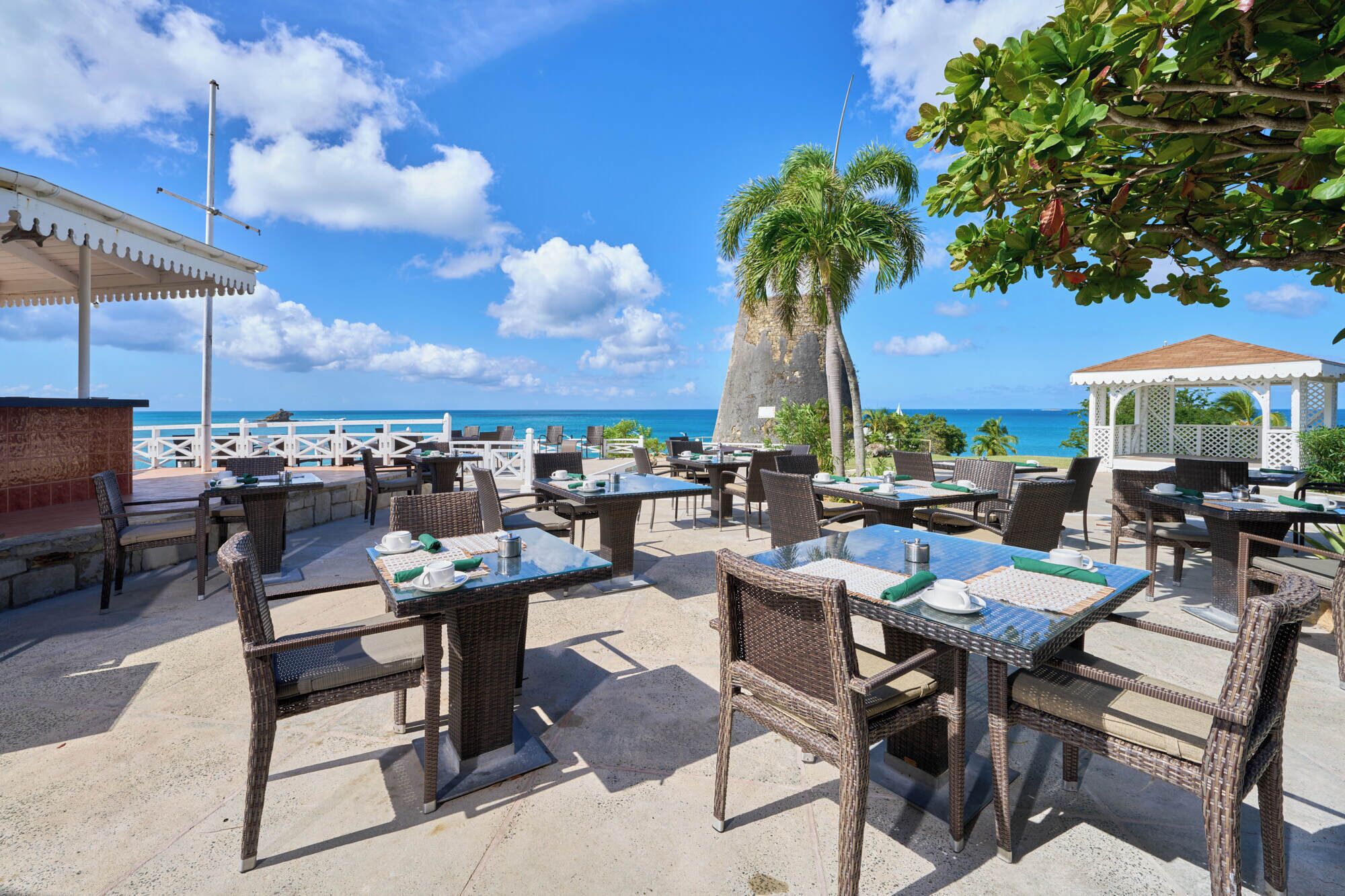 Un espace de restauration en plein air, doté de mobilier et de tables en osier, offrant une vue sur une plage et l’océan.