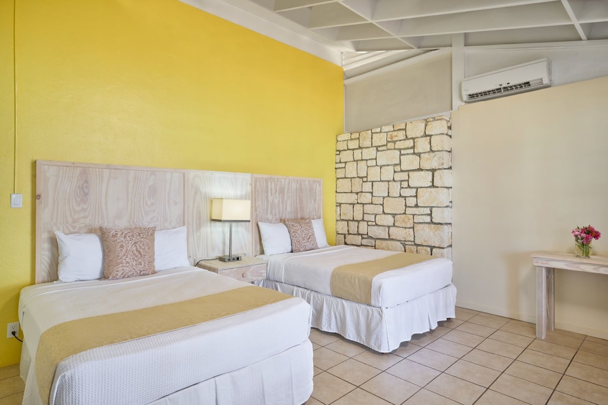 A hotel room with two beds, a stone wall, an air conditioning unit, and a small table with flowers.