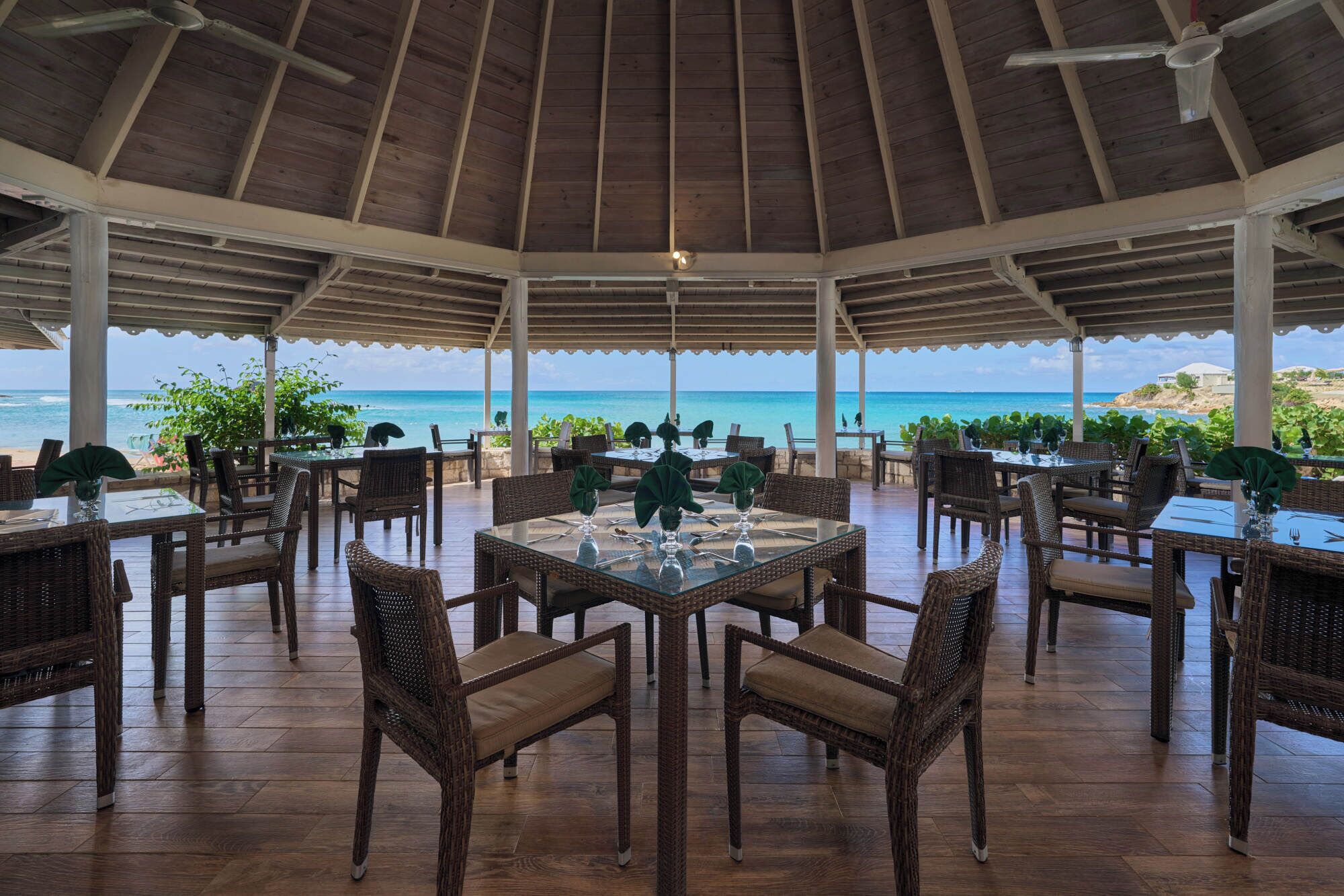 An outdoor dining area with wicker furniture and glass tables, overlooking a beach.