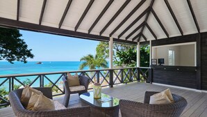 A covered outdoor seating area with wicker furniture and a glass table, overlooking a clear blue sea and a rocky island. - Hawksbill Resort Antigua (St. John's)