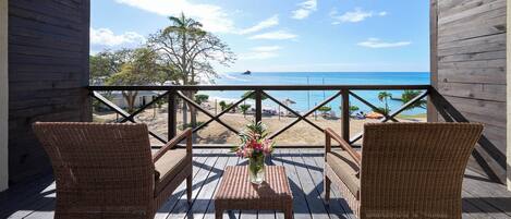 A wooden deck with wicker chairs and a table, overlooking a beach with palm trees and the ocean.