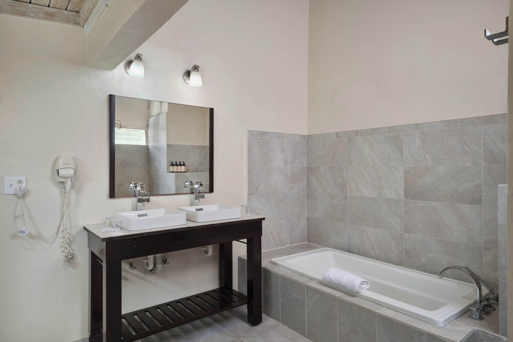 Ein modernes Badezimmer mit zwei Waschbecken, einem großen Spiegel, einem Waschtisch, einer Badewanne und einem Föhn an der Wand.