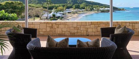 A covered outdoor seating area with wicker furniture and cushions, overlooking a beach and ocean.