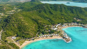 An aerial view of a resort with a beach, clear turquoise waters, and lush green hills. - Hawksbill Resort Antigua (St. John's)