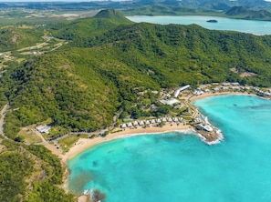 An aerial view of a resort with a beach, clear turquoise waters, and lush green hills. - Hawksbill Resort Antigua (St. John's)