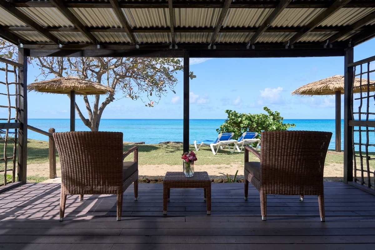 A covered outdoor seating area with wicker chairs and a table, overlooking a beach with lounge chairs and a clear view of the ocean.