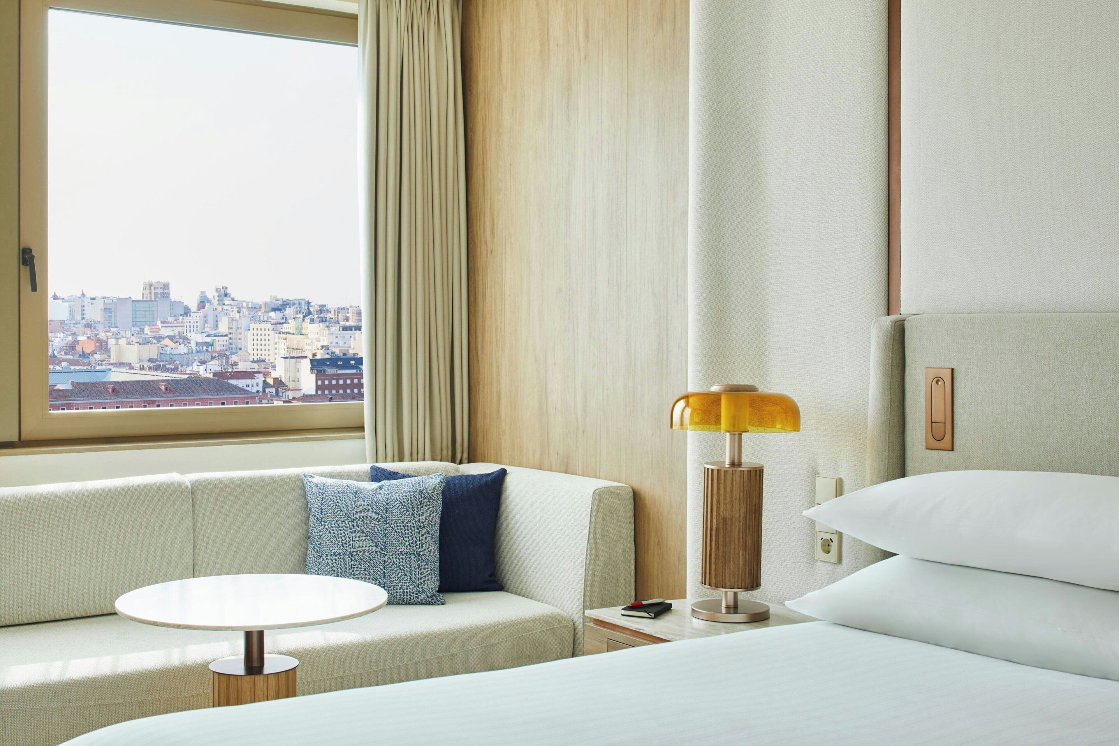 Quarto de hotel com sofá, uma mesinha redonda, cama com roupa de cama branca, abajur e vista para a paisagem urbana pela janela.