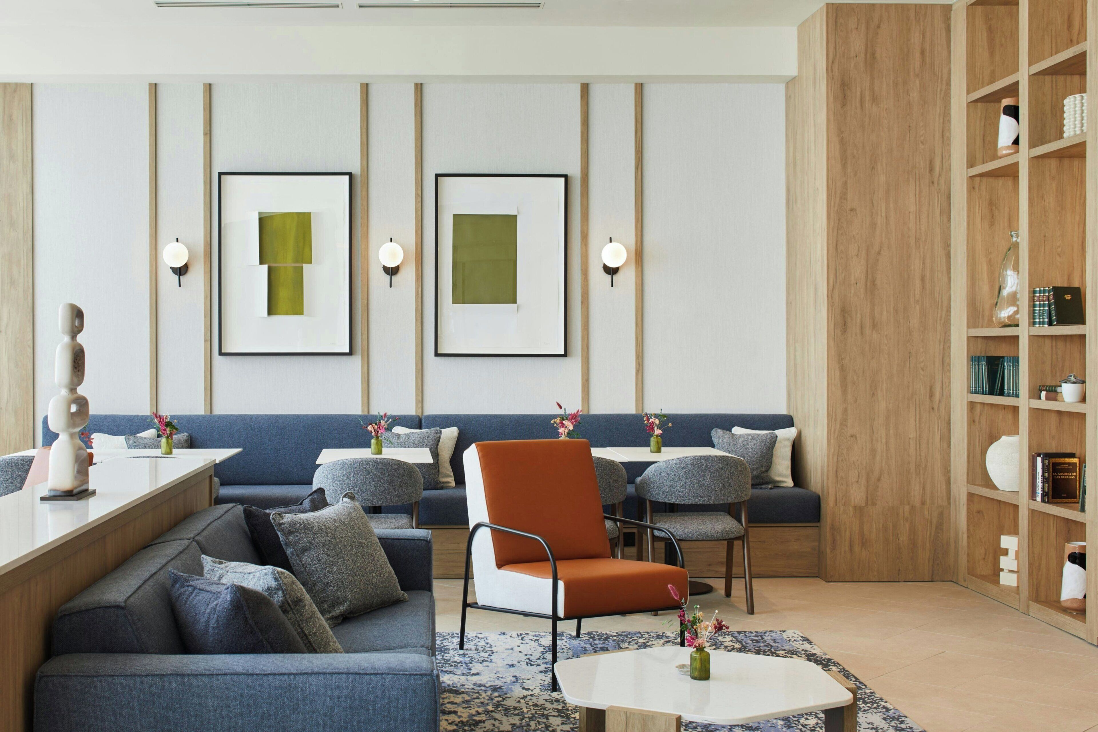 A modern lounge area with seating arrangements, framed artwork on the wall, and a wooden bookshelf.