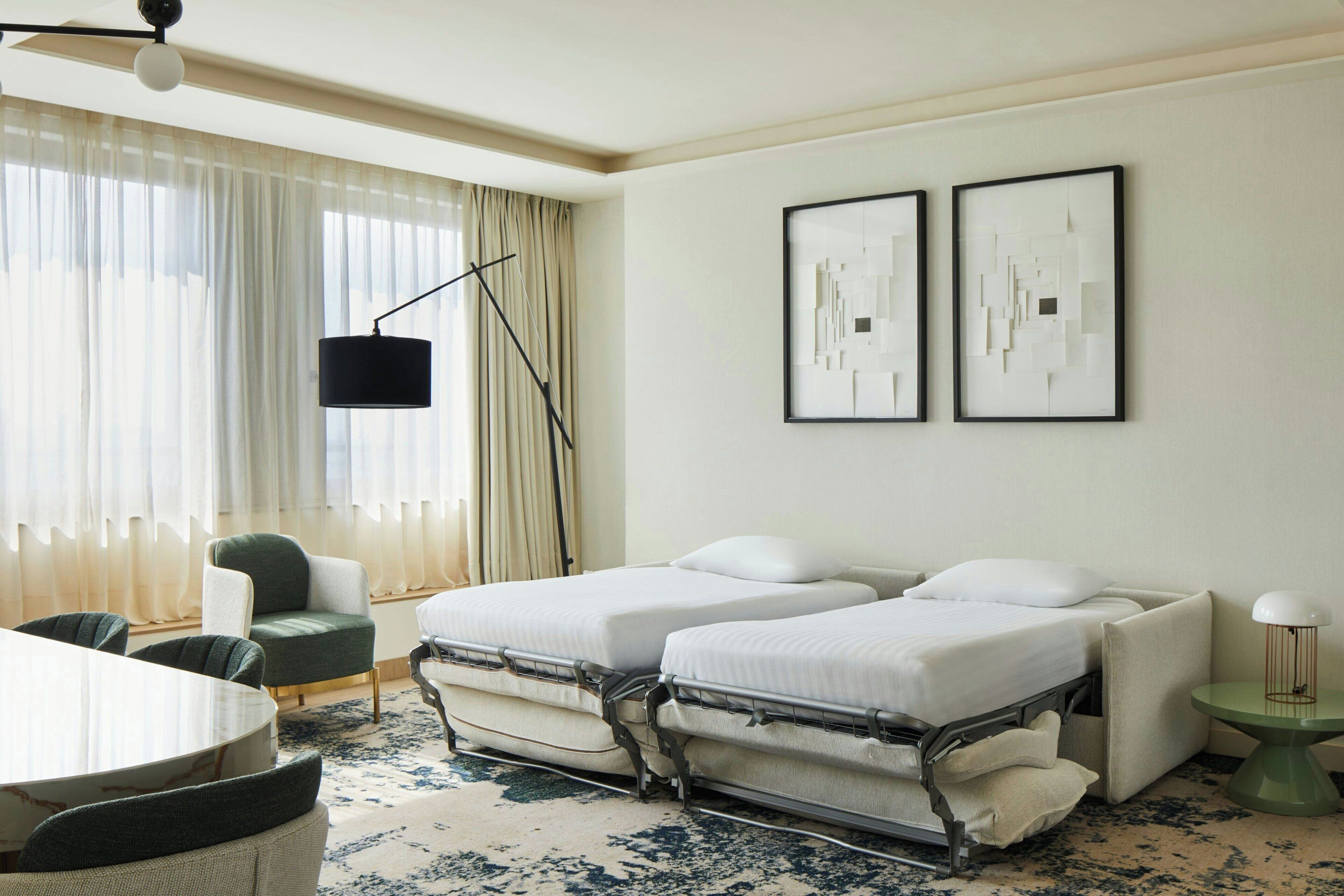 A hotel room with two beds, a dining table, a chair, a floor lamp, and two framed abstract artworks on the wall.