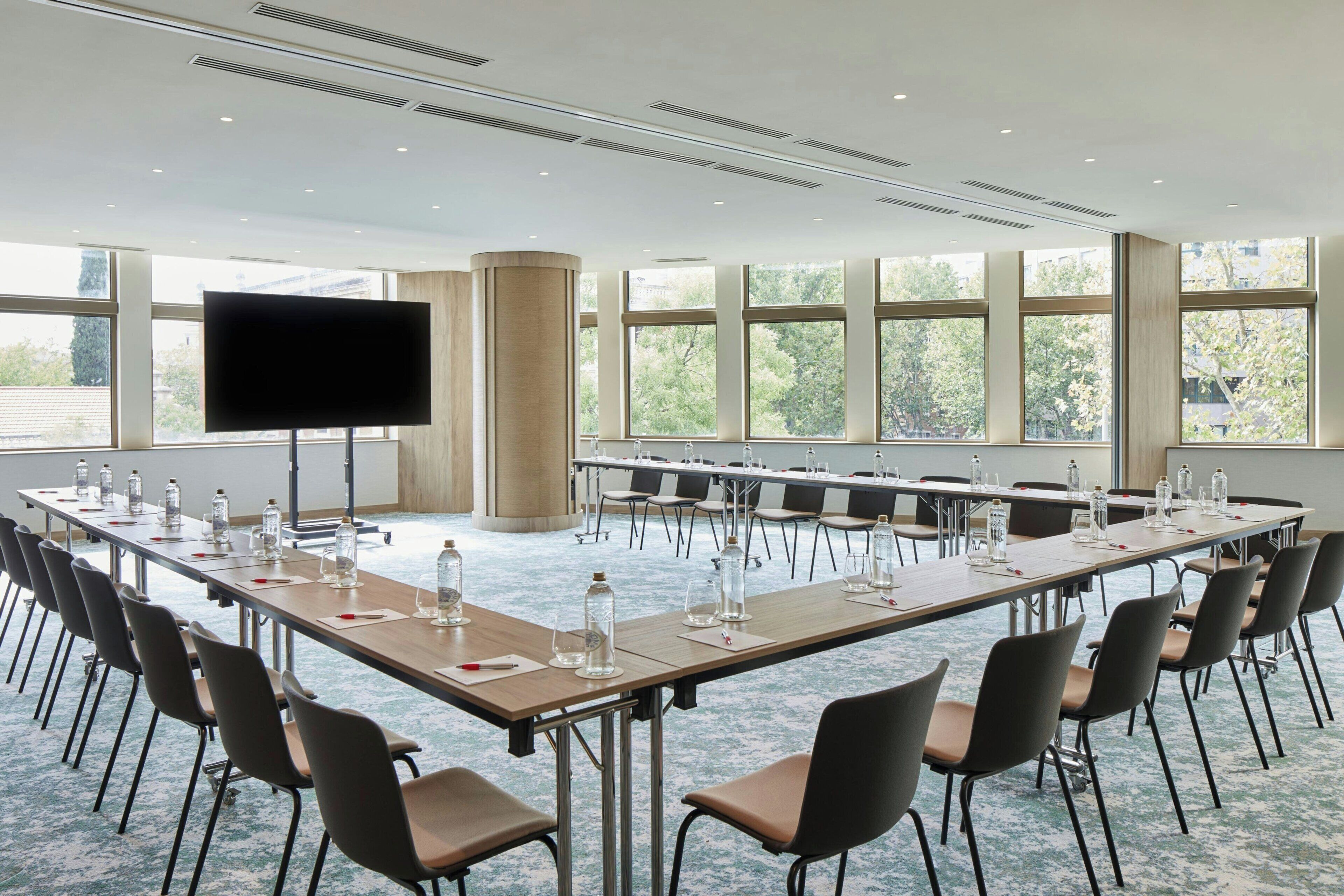 Un salón de conferencias con una larga mesa, sillas, una pantalla grande y ventanas que dan a vistas de árboles.