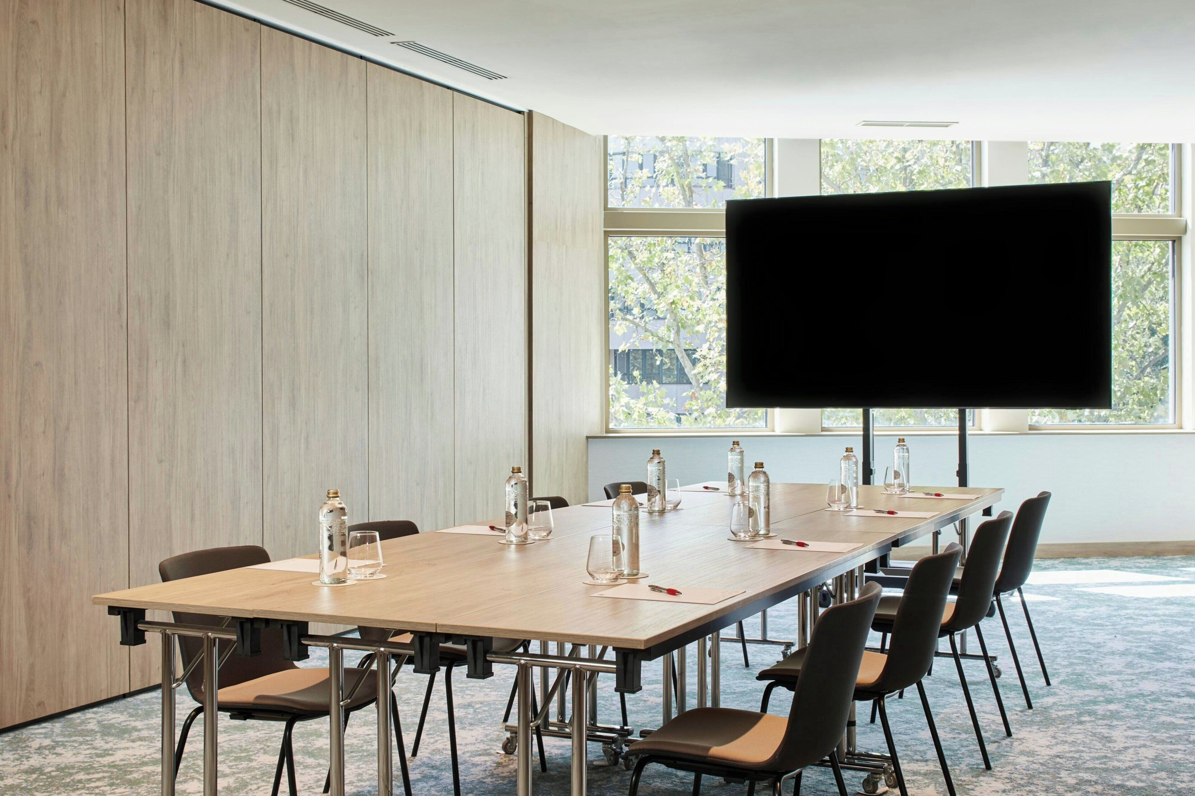 A conference room with a long table, chairs, a large screen, and a window with a view of trees.