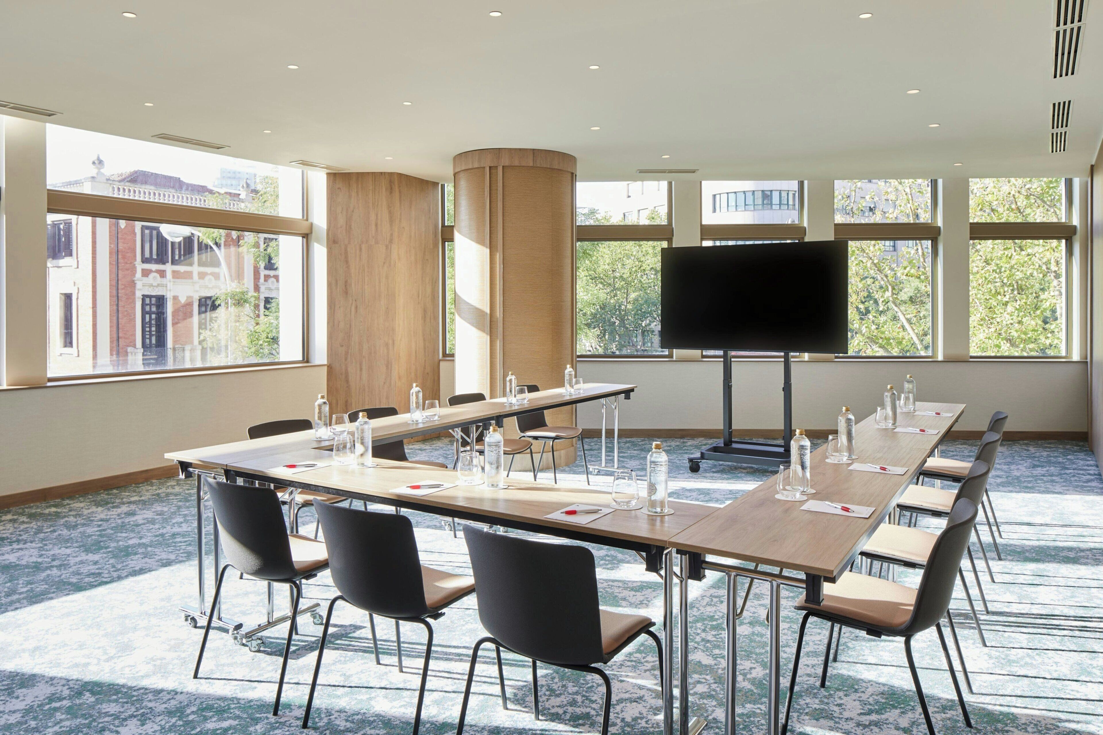 Un salón de conferencias con una larga mesa, sillas, un televisor de pantalla plana y amplias ventanas que ofrecen vistas a edificios y árboles.