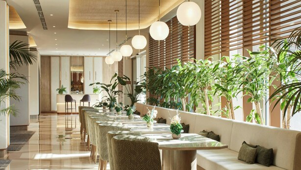 Un amplio y luminoso vestíbulo de hotel con una larga mesa de comedor, asientos cómodos y abundante vegetación interior.