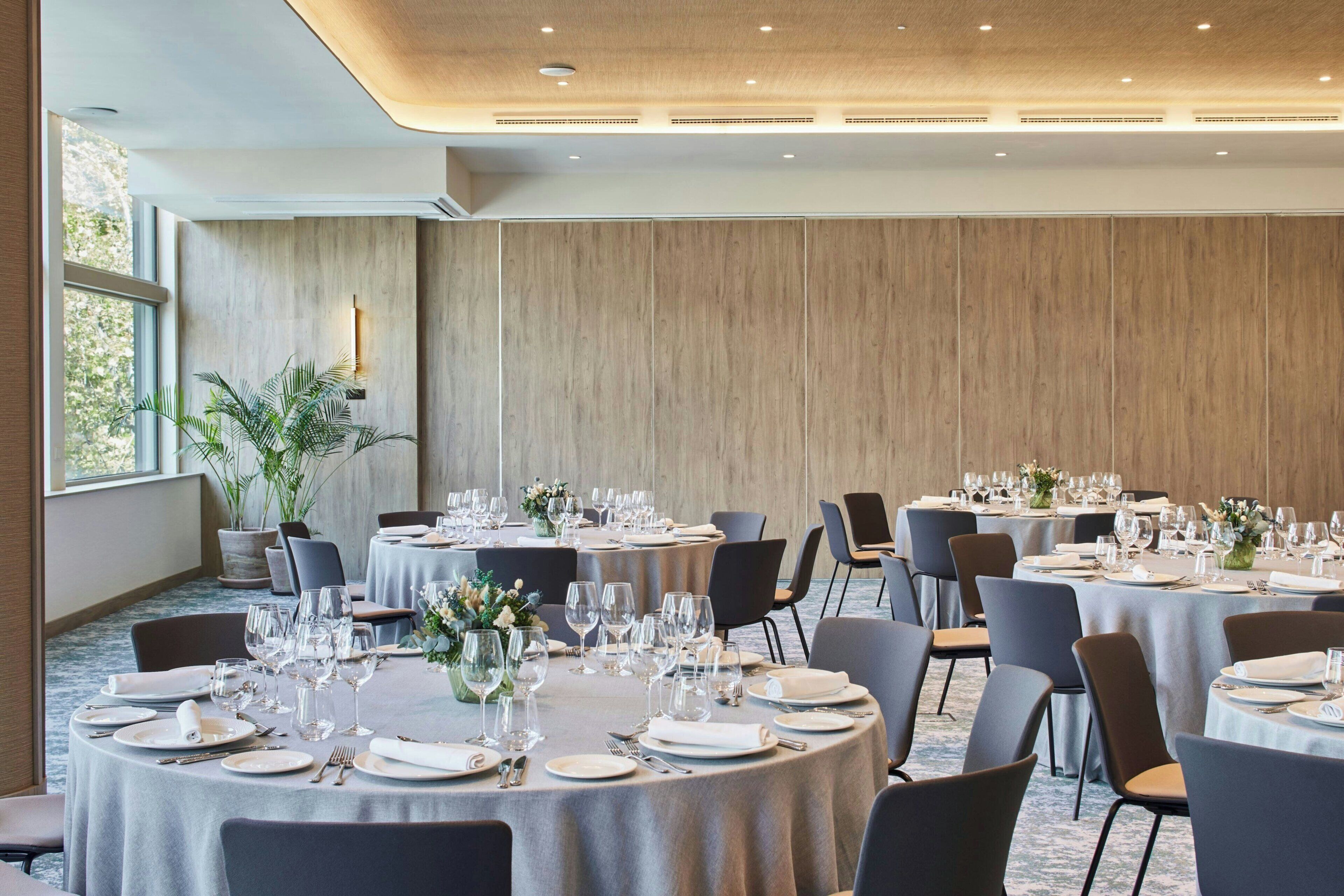 Un salón de conferencias con mesas redondas dispuestas para un evento formal, equipado con sillas, platos, vasos y cubiertos.