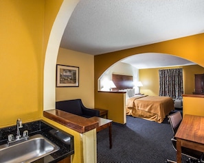 A hotel room with a kitchenette, a bed with a headboard, a desk, and a chair.