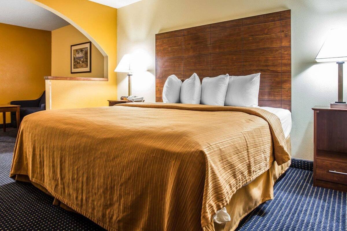 A hotel room with a large bed, wooden headboard, two bedside tables with lamps, a small desk, and a chair.