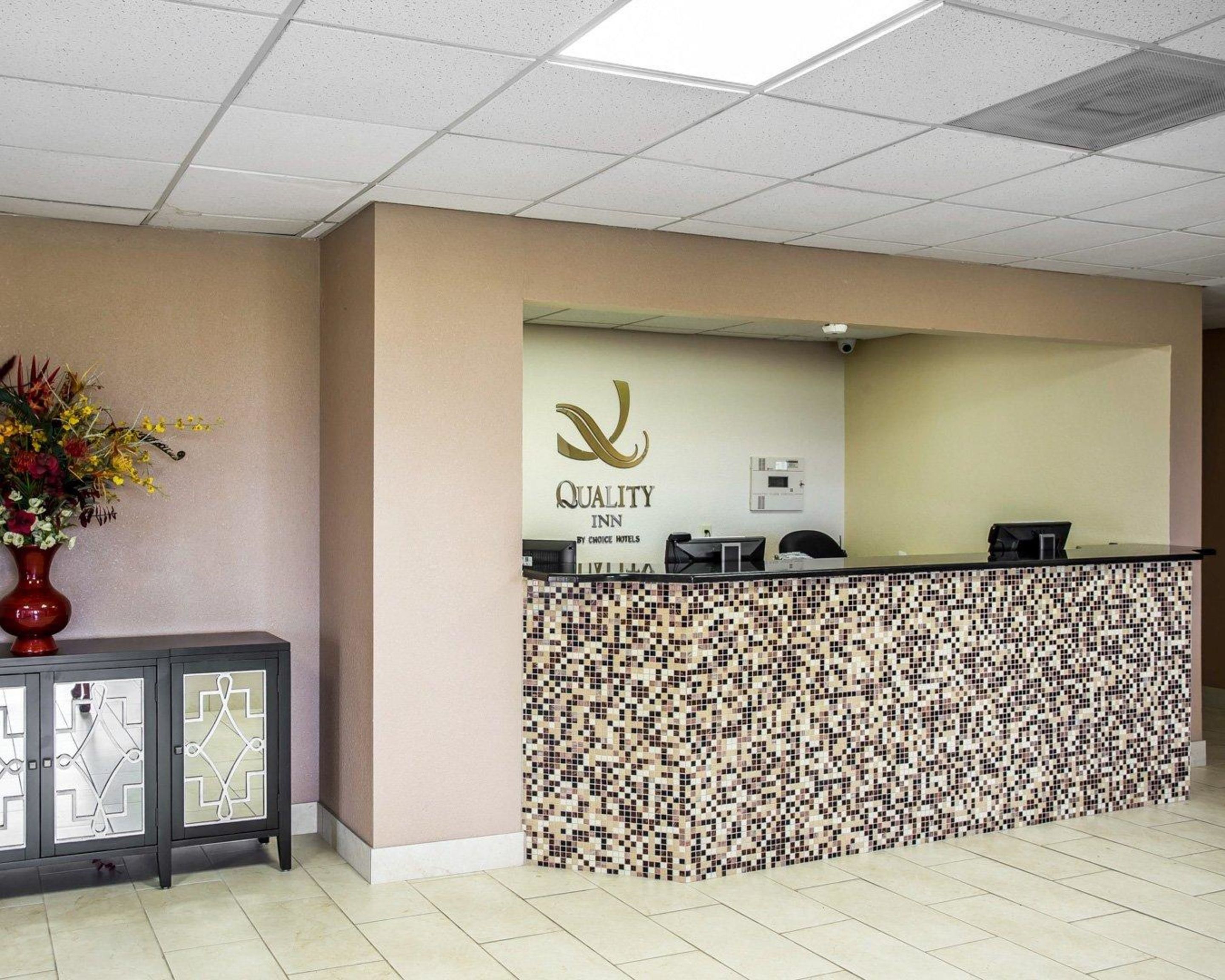 Un mostrador de recepción con diseño de mosaico, el logotipo de 'Quality Inn by Choice Hotels' y un jarrón con flores a la izquierda.
