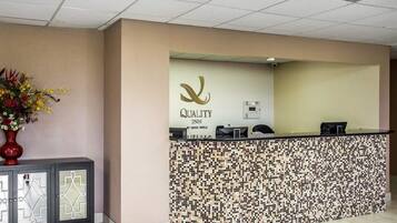 Un mostrador de recepción con diseño de mosaico, el logotipo de 'Quality Inn by Choice Hotels' y un jarrón con flores a la izquierda.