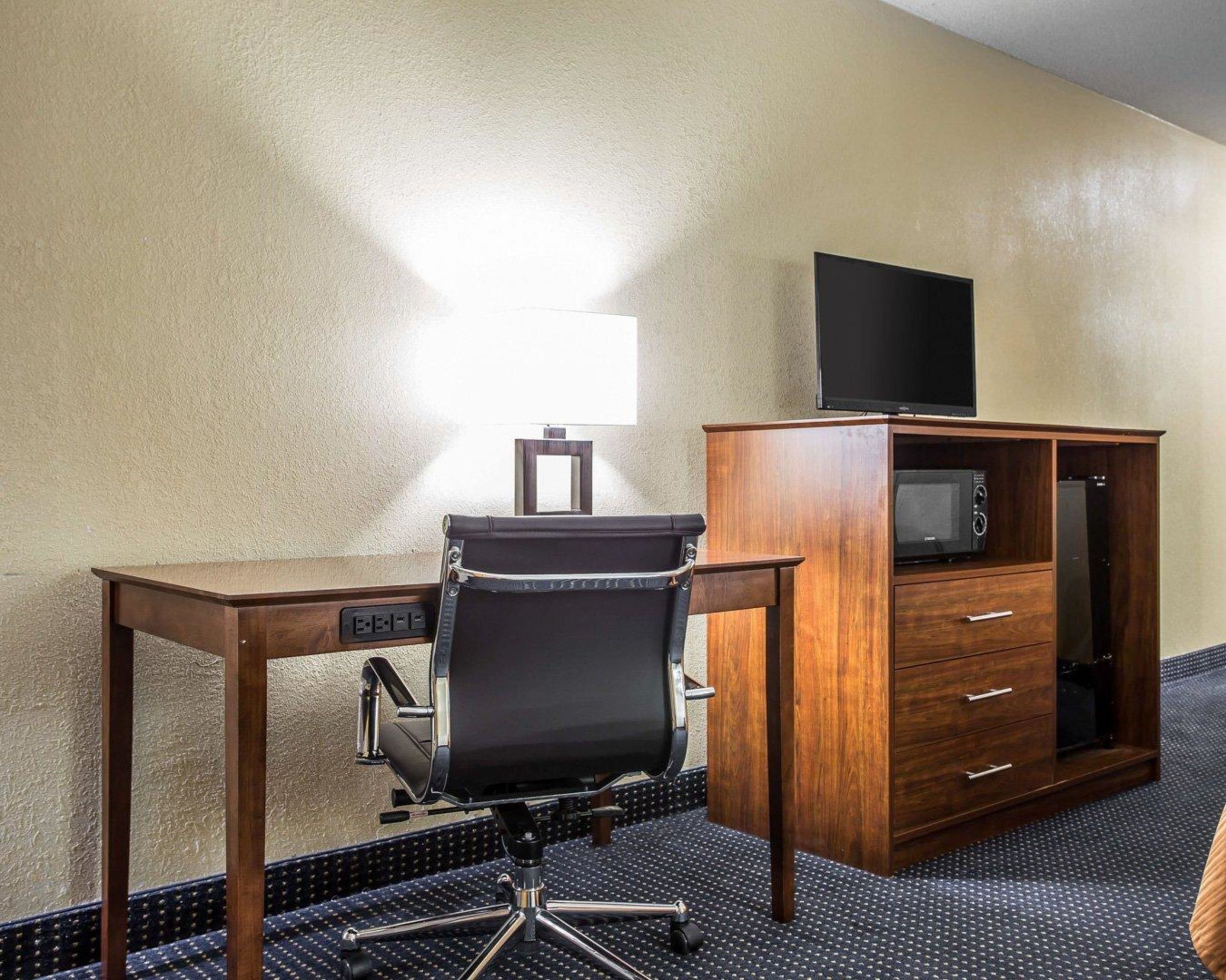 A hotel room with a wooden desk, a black office chair, a flat-screen TV on a cabinet, a microwave, and a lamp.