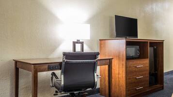 A hotel room with a wooden desk, a black office chair, a flat-screen TV on a cabinet, a microwave, and a lamp.