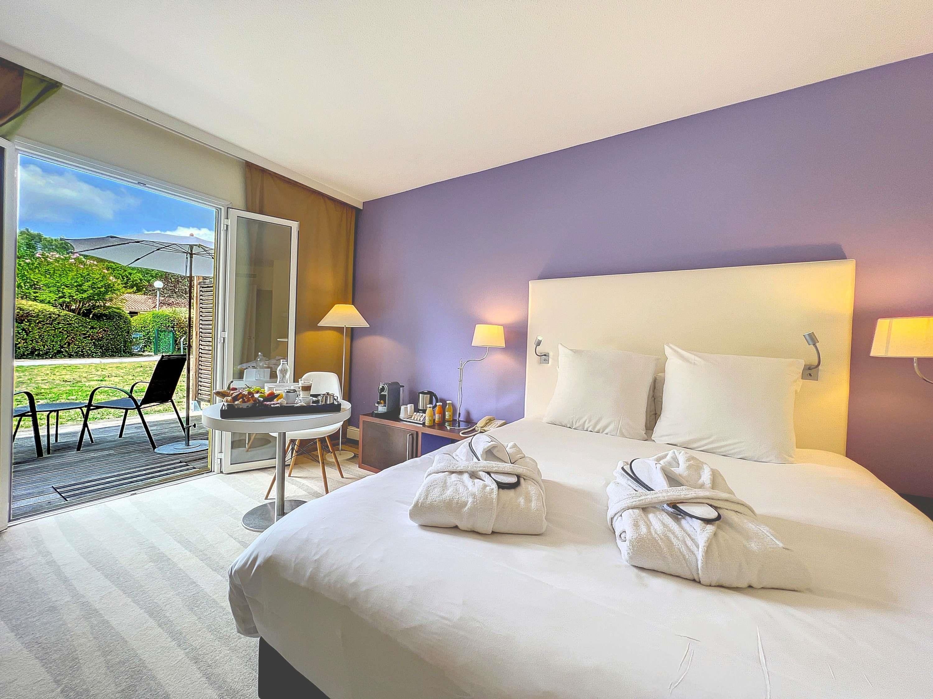A hotel room with a bed, a desk with a chair, a table with breakfast items, and a view of the outdoors.