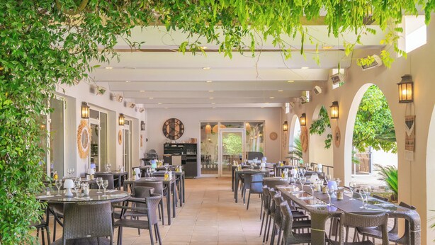 An outdoor dining area with wicker tables and chairs, surrounded by arches and greenery.