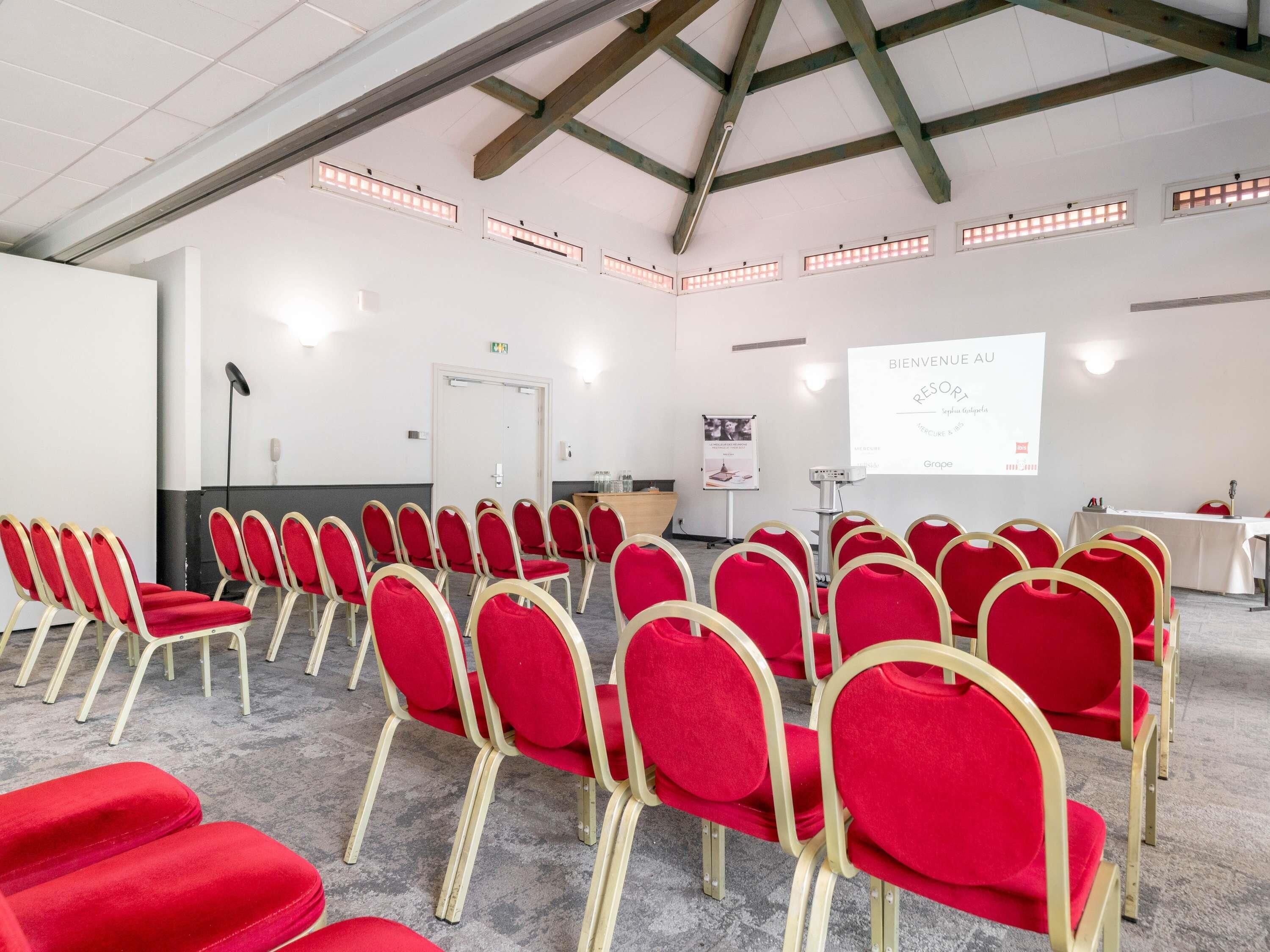 Une salle de conférence avec des rangées de chaises rouges, un écran de projection et un pupitre.