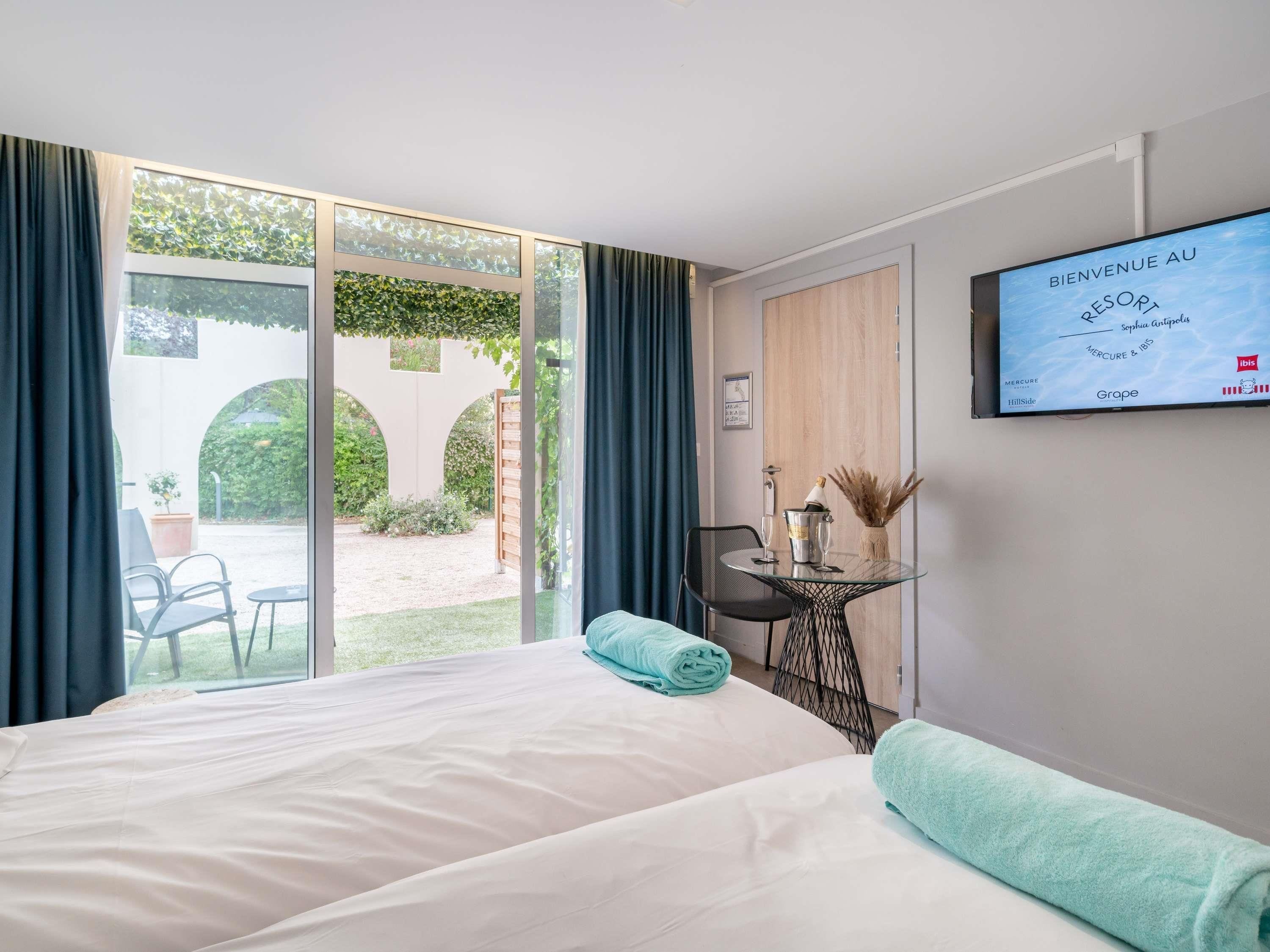 Une chambre d’hôtel avec un lit, un bureau et une chaise, un téléviseur fixé au mur et une vue sur le jardin.