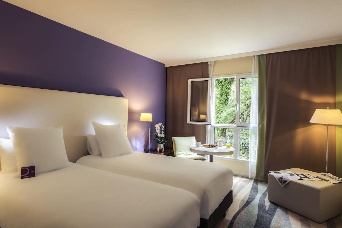 Hotellihuone, jossa on kaksi sänkyä, pieni pöytä lampun kanssa, ikkuna verhoilla ja näkymä vehreään maisemaan.