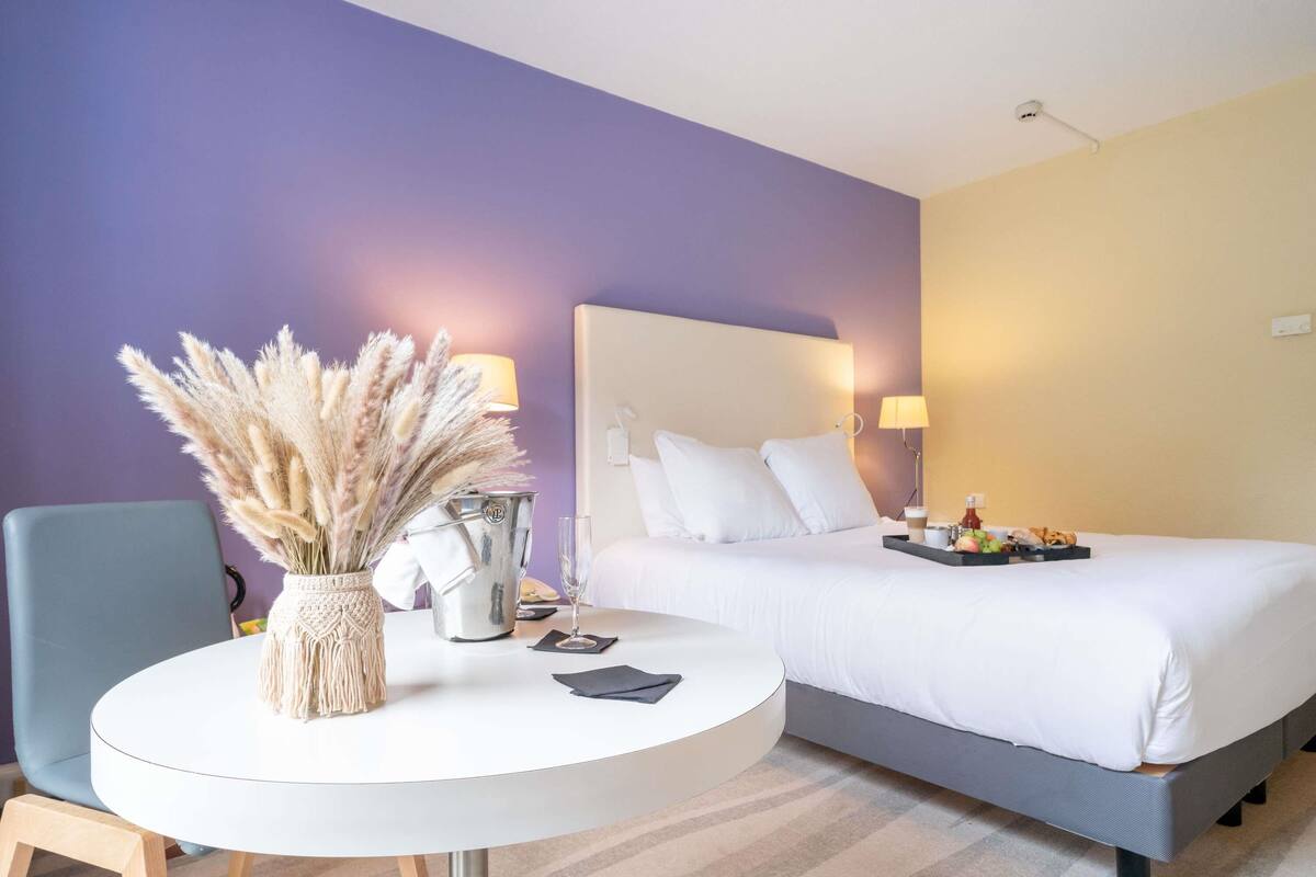 Ein Hotelzimmer mit einem Bett, einem runden Tisch mit einer Vase mit Pampasgras, einem Eimer und einem Tablett mit Essen.