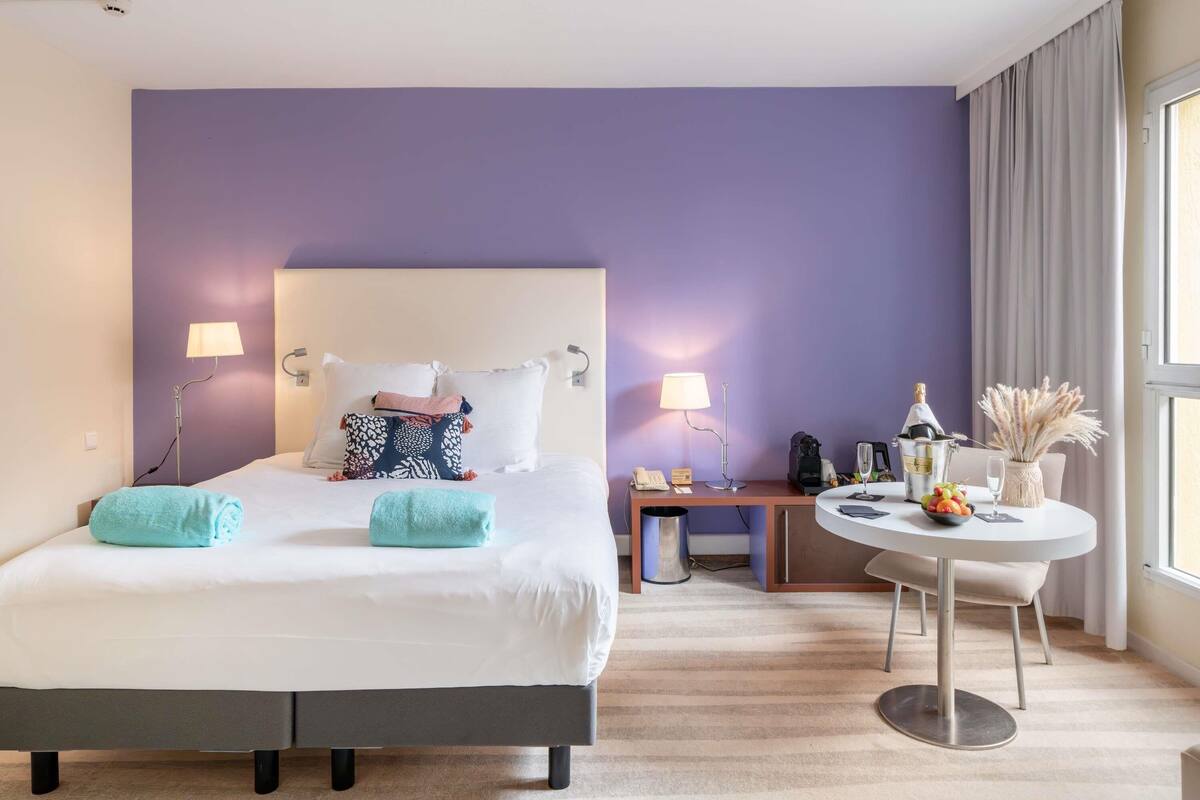 Hotelkamer met een bed, bedlampjes, een klein tafeltje met een fles, een vaas met gedroogde bloemen en een raam met gordijnen.