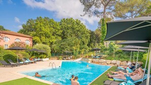 A swimming pool with lounge chairs and umbrellas, surrounded by greenery and a building.