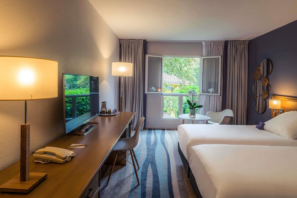 Une chambre d’hôtel équipée d’un bureau, de deux lits, d’une télévision et d’une fenêtre avec des rideaux.