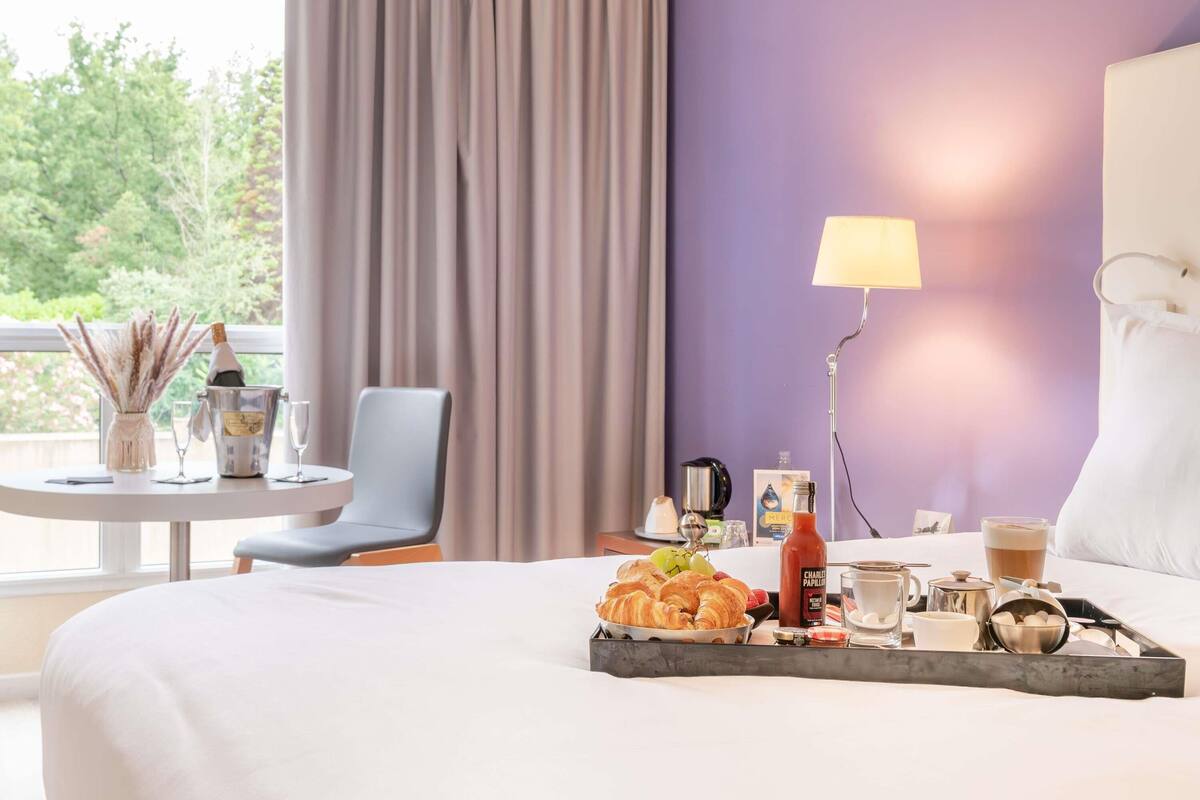 Een hotelkamer met een bed, een tafel met ontbijtartikelen, een stoel, een lamp en uitzicht op groen.