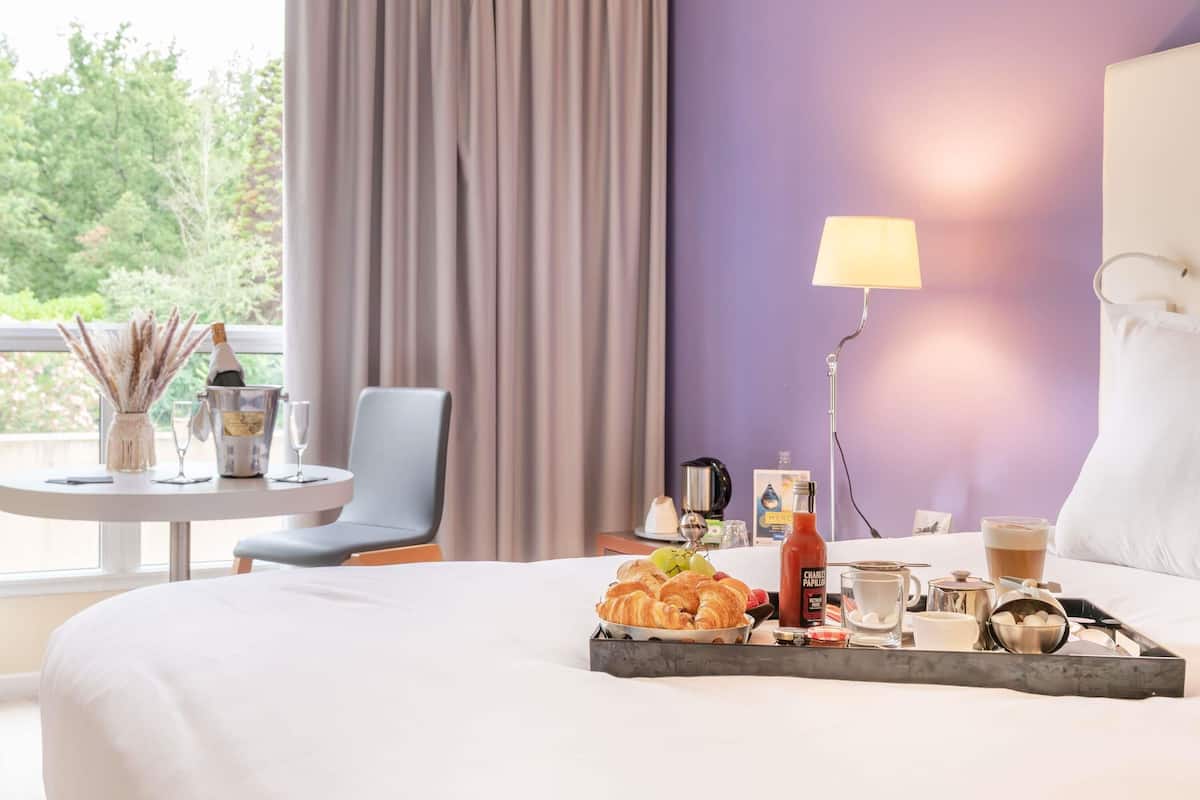 Ein Hotelzimmer mit Bett, Tisch mit Frühstücksgebäck, einem Stuhl, einer Lampe und Blick auf Grünfläche.