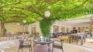 An indoor dining area with round tables and wicker chairs, surrounded by lush greenery and hanging light fixtures.