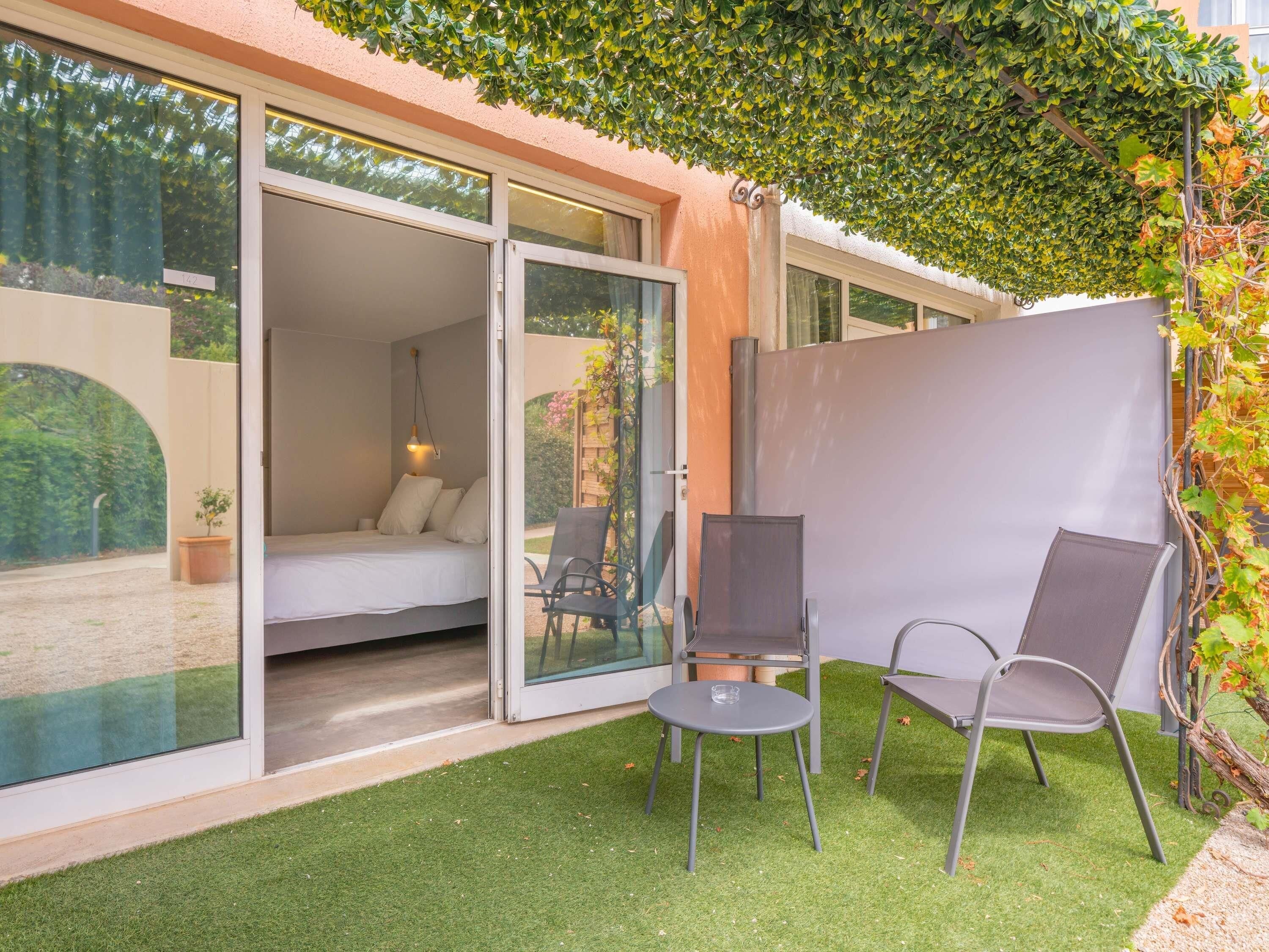A modern room with a bed, a chair, a table, and a glass door leading to an outdoor area with a view of greenery.