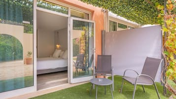 A modern room with a bed, a chair, a table, and a glass door leading to an outdoor area with a view of greenery.