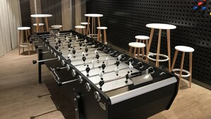 Une table de baby-foot dans une pièce avec un mur noir perforé, des tabourets en bois et des luminaires suspendus.