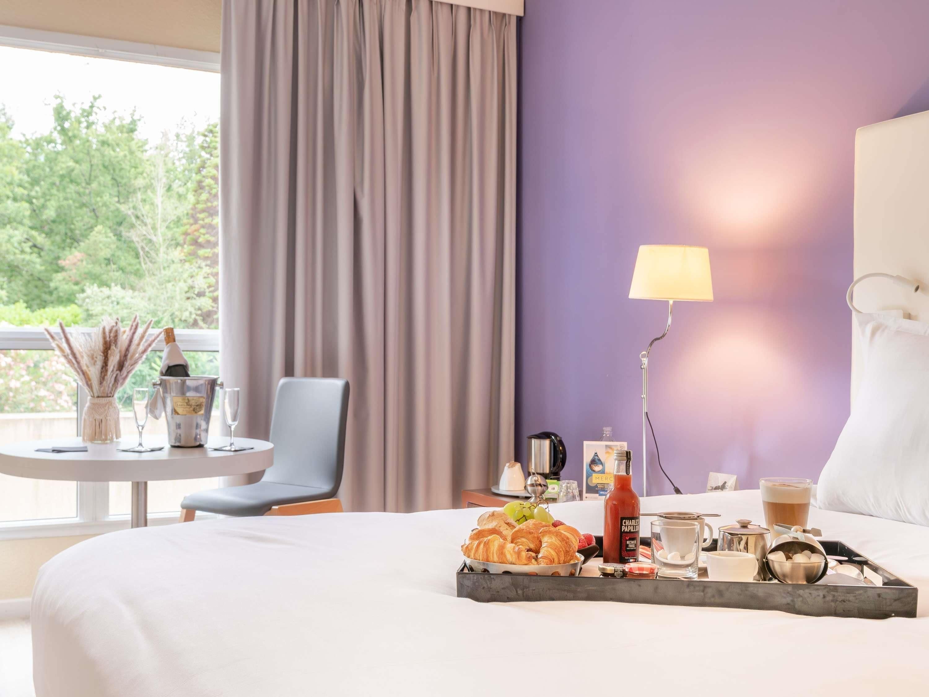A hotel room with a bed, a table with breakfast items, a chair, a lamp, and a view of greenery outside.