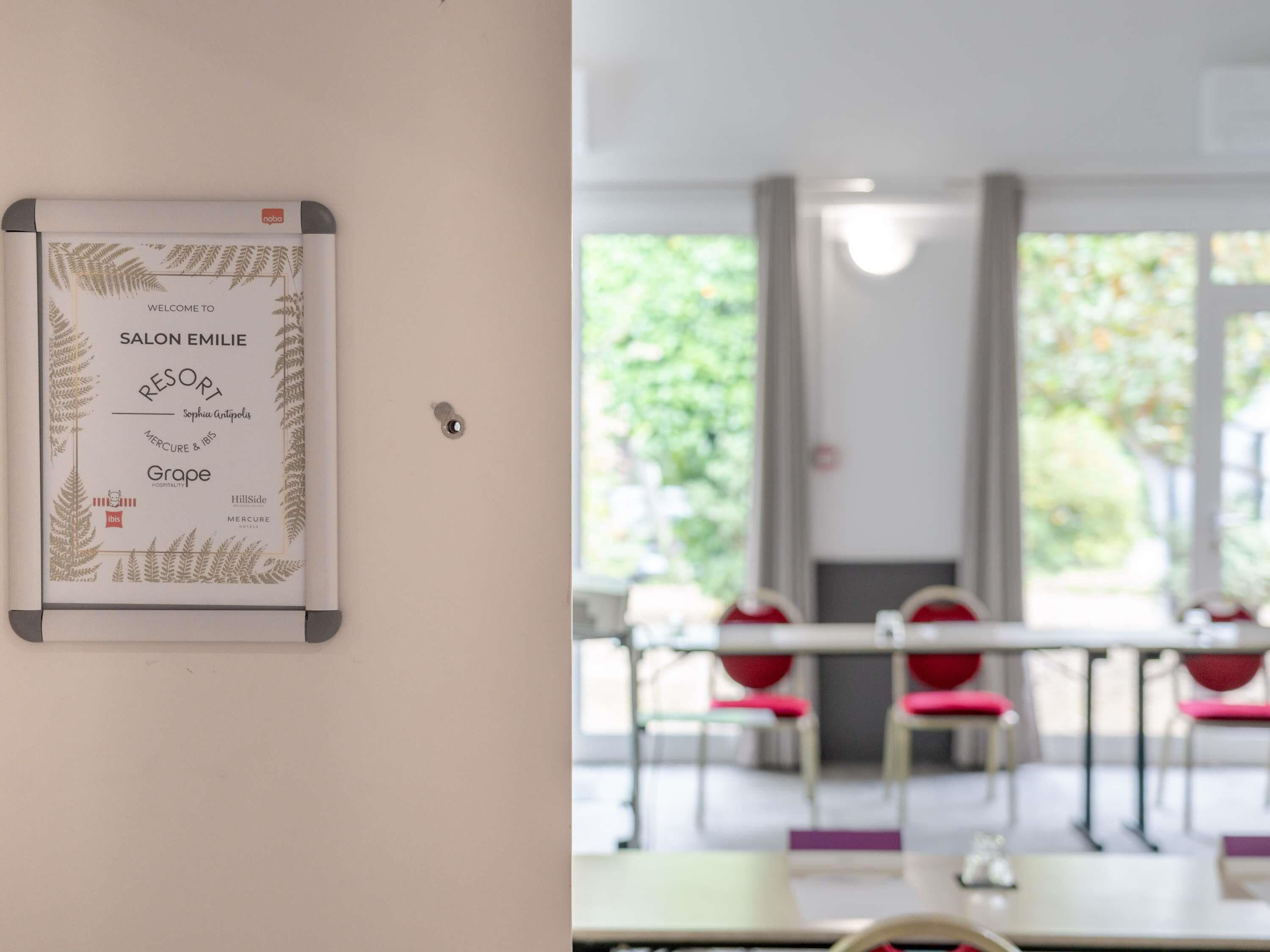 A framed welcome sign for Salon Emilie Resort, a room with a view of greenery, and red chairs arranged around tables.