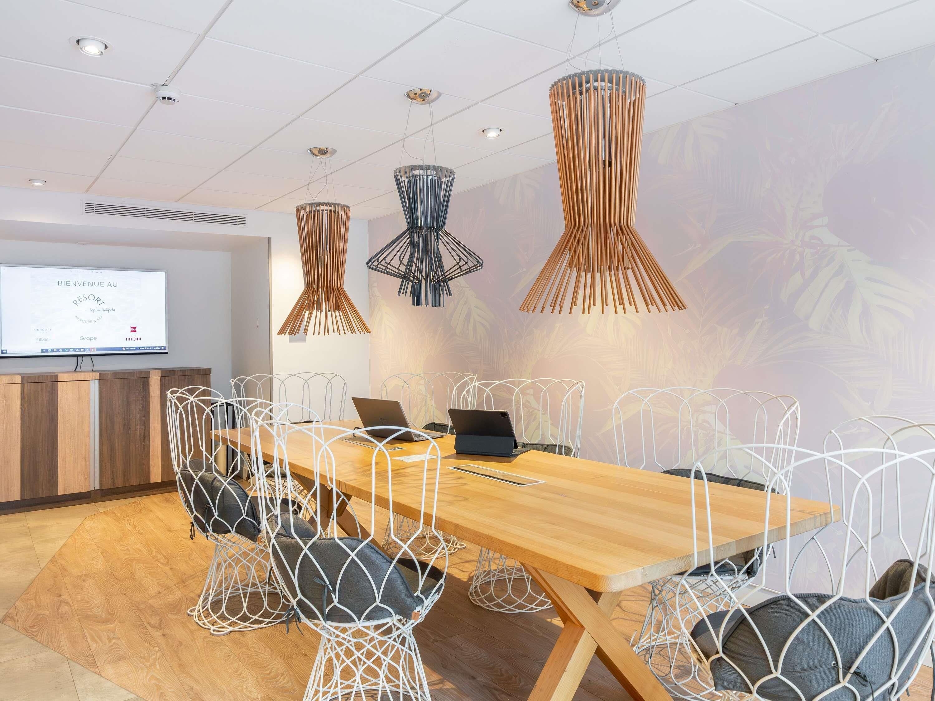 A conference room with a long wooden table, white wireframe chairs, and three hanging pendant lights.