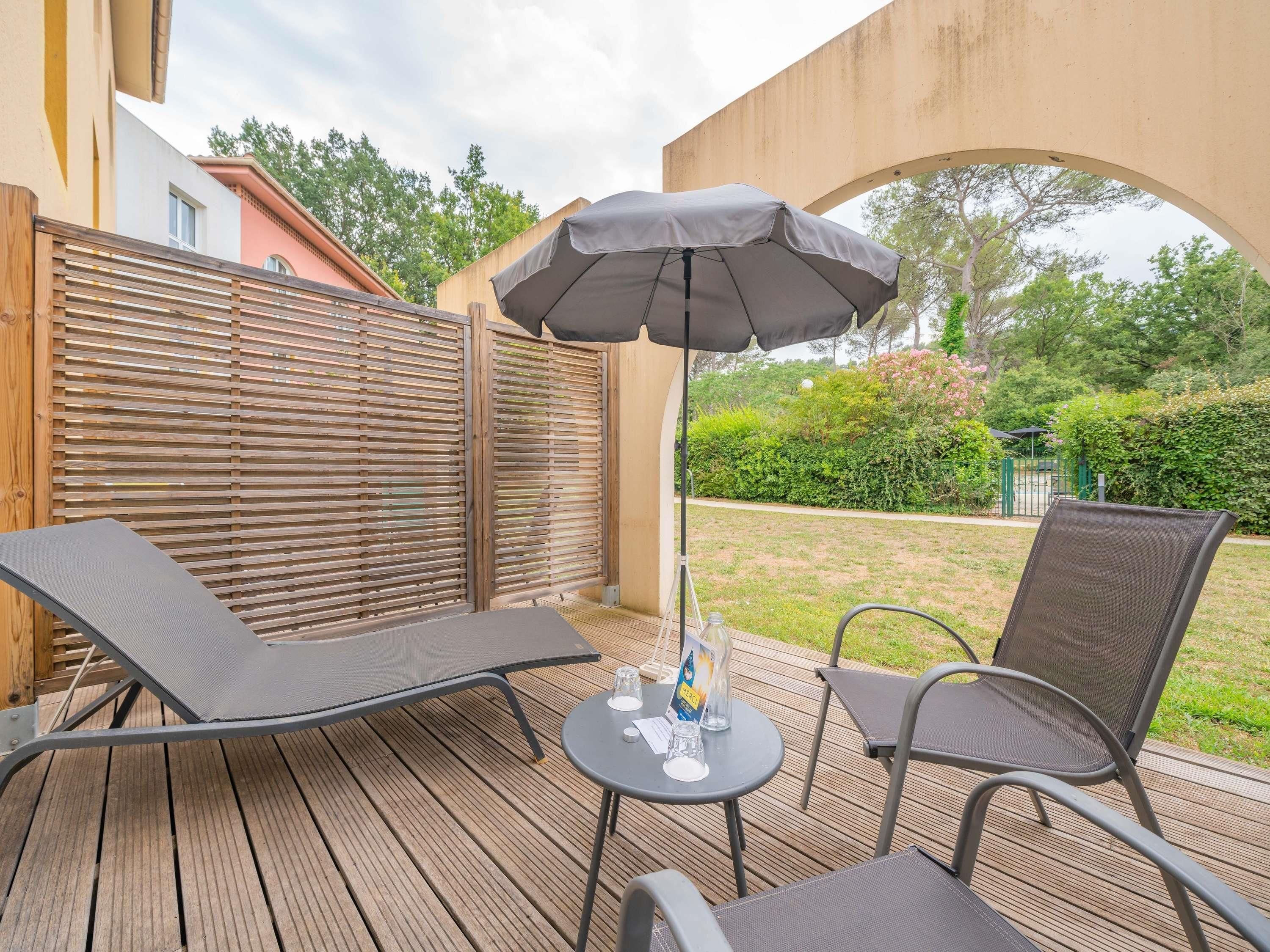 A wooden deck with two lounge chairs and a table, an umbrella, and a view of a garden.