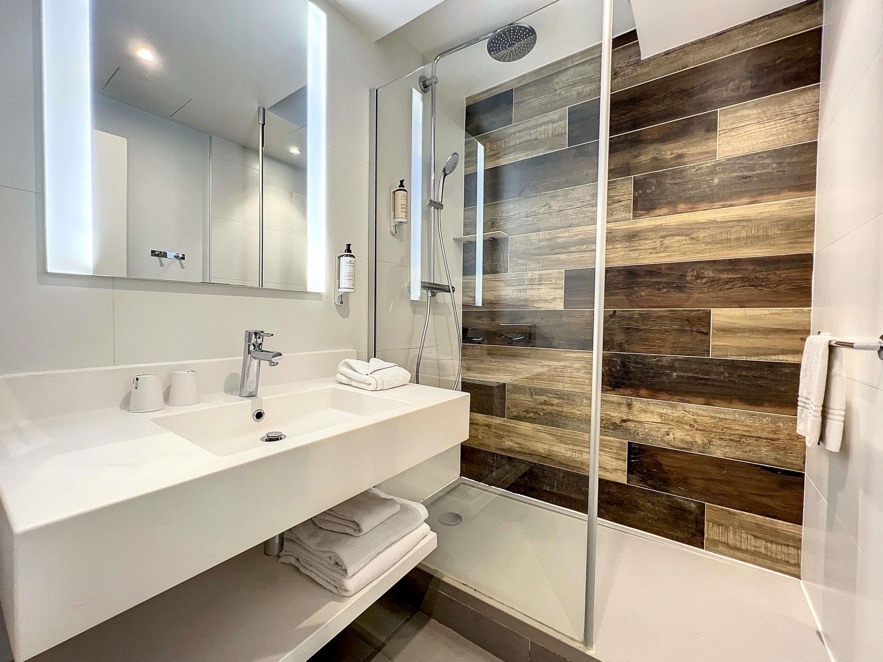 A modern bathroom with a white sink, a mirror, a glass shower enclosure, and a wooden wall design.