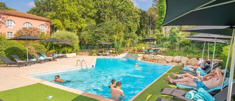 A swimming pool with lounge chairs and umbrellas, surrounded by greenery and a building.
