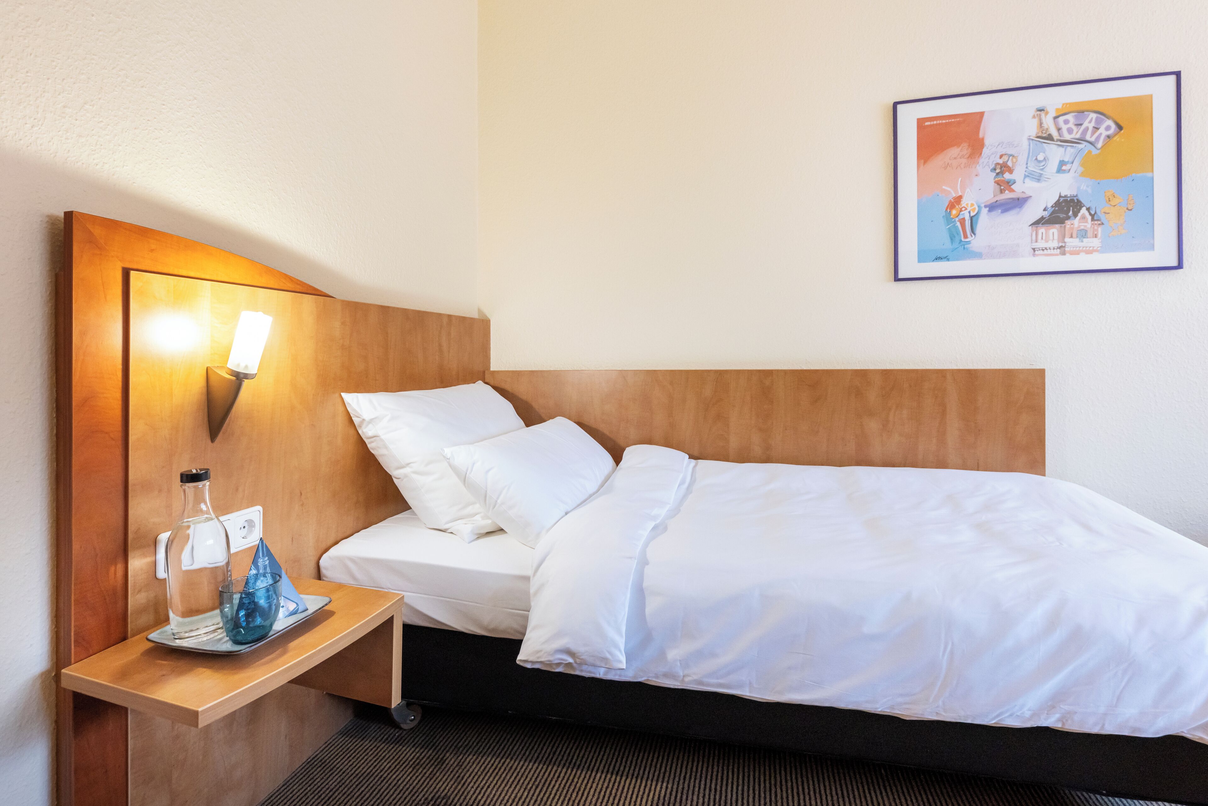 Ein Hotelzimmer mit einem Bett, einem Nachttisch mit Lampe, einem Glas Wasser und einem gerahmten Bild an der Wand.