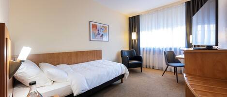 A hotel room with a bed, two chairs, a desk, a lamp, a picture on the wall, and a window with curtains.