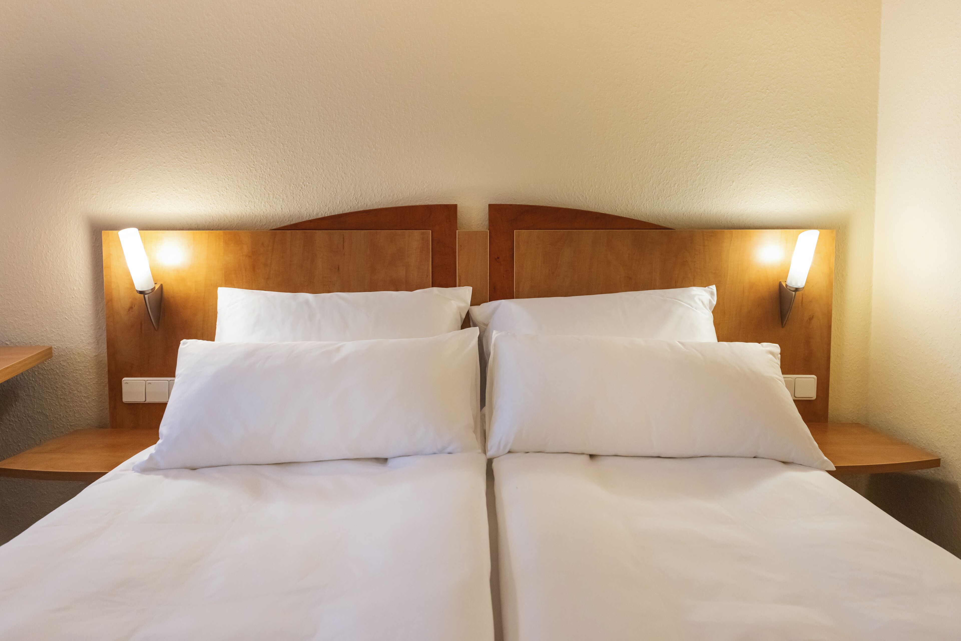 Un lit double avec du linge de lit blanc, deux tables de chevet et des lampes fixées au mur.