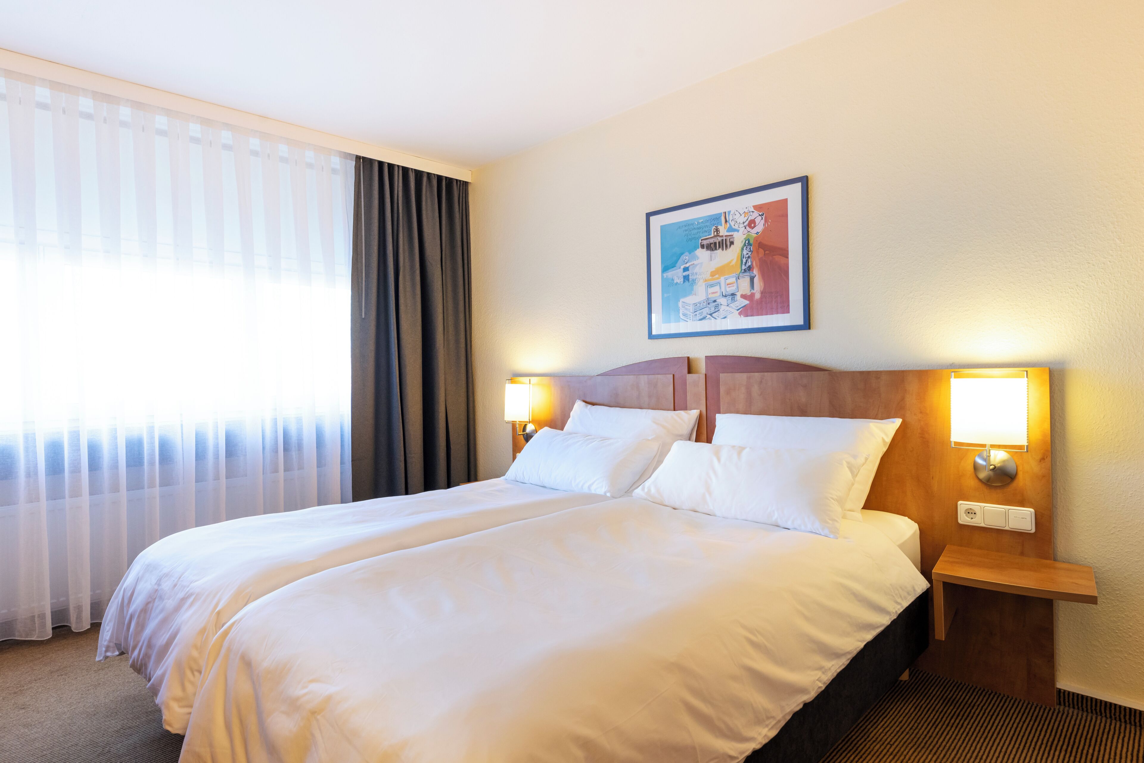Una camera d'albergo con un letto matrimoniale, due lampade ai comodini, un quadro appeso al muro e una finestra con tende.